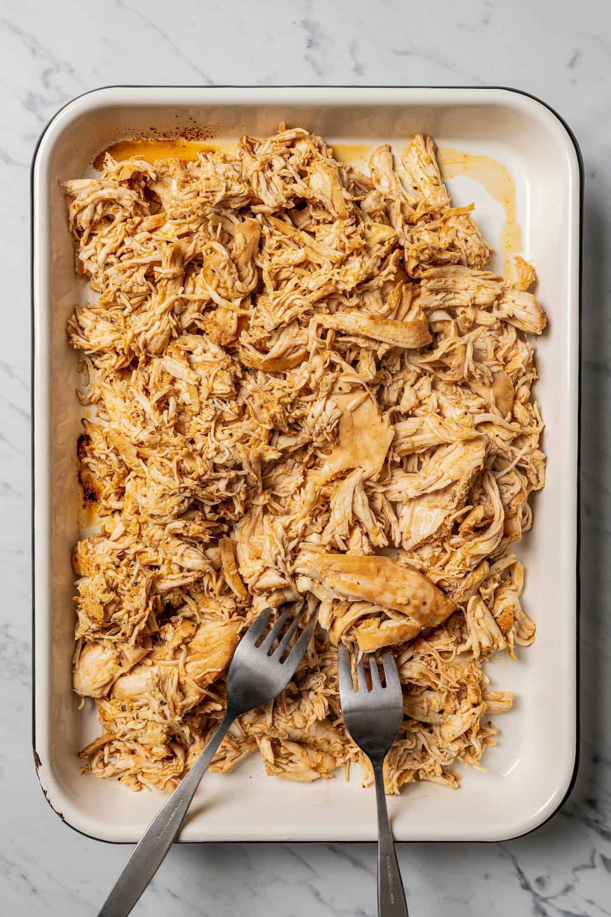 Shredded chicken in a rimmed baking tray with two forks.