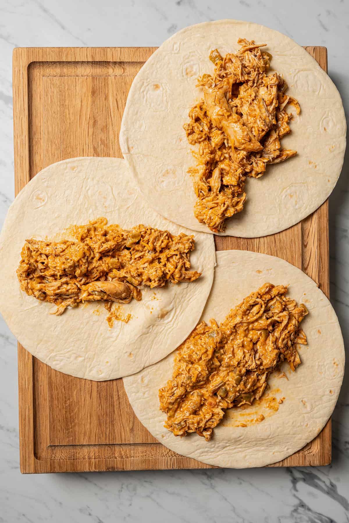 Overhead view of three flour tortillas topped with chicken filling on a wooden cutting board.