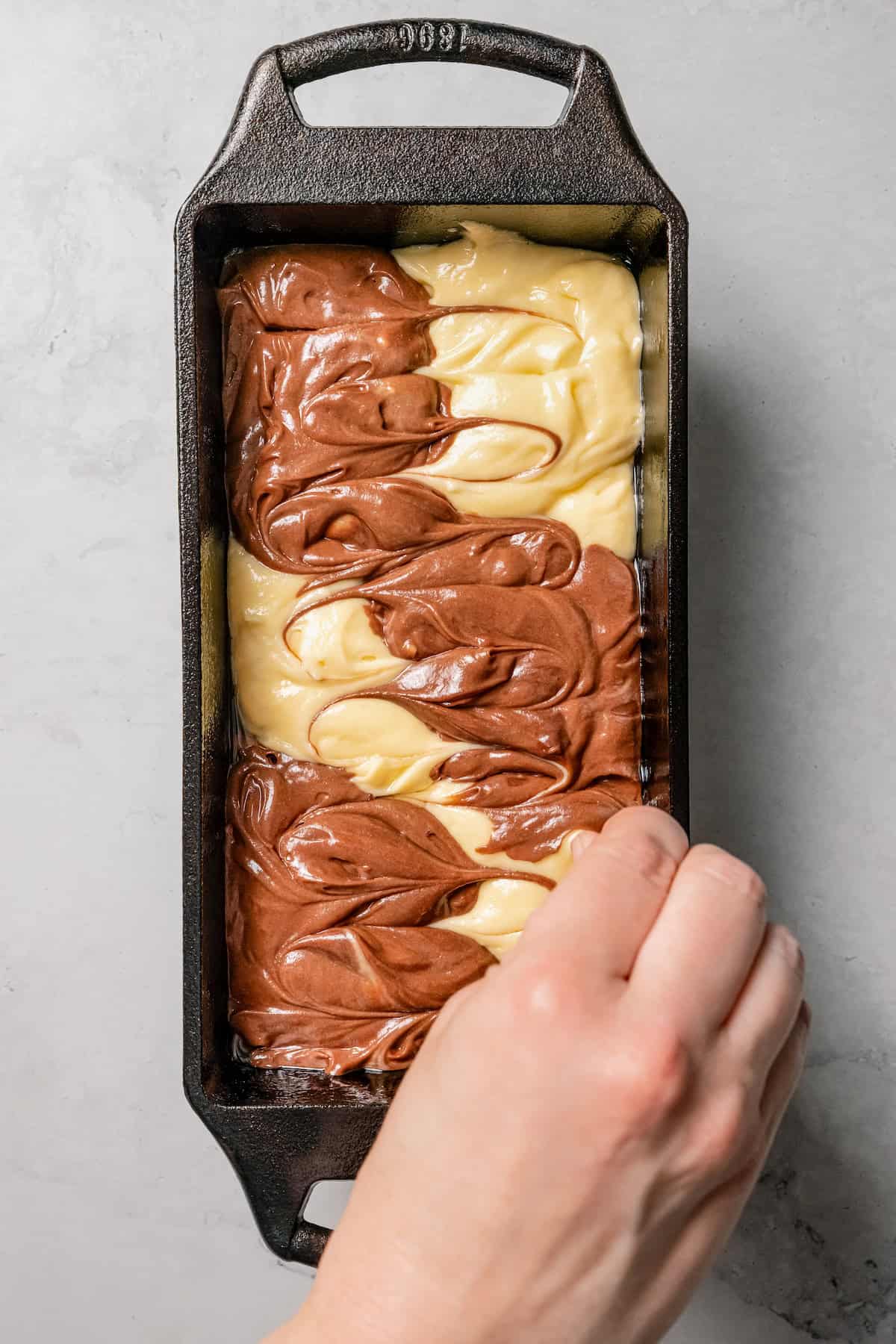 A hand uses a toothpick to swirl chocolate and vanilla cake batter together in a loaf pan.
