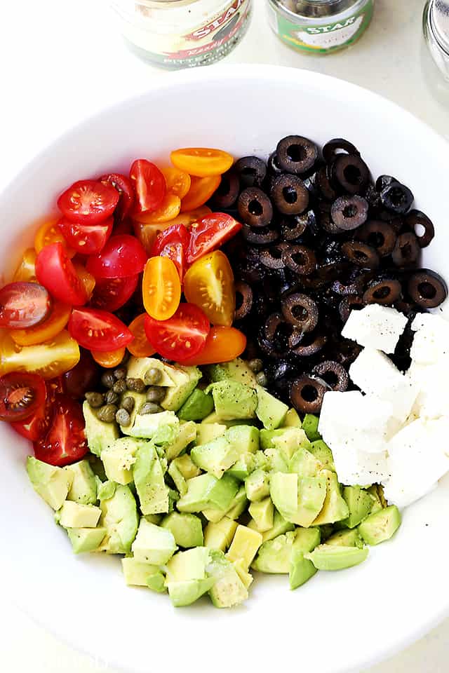 A white big bowl filled with diced avocado, sliced olives, halved cherry tomatoes, feta cheese cubes, and capers.
