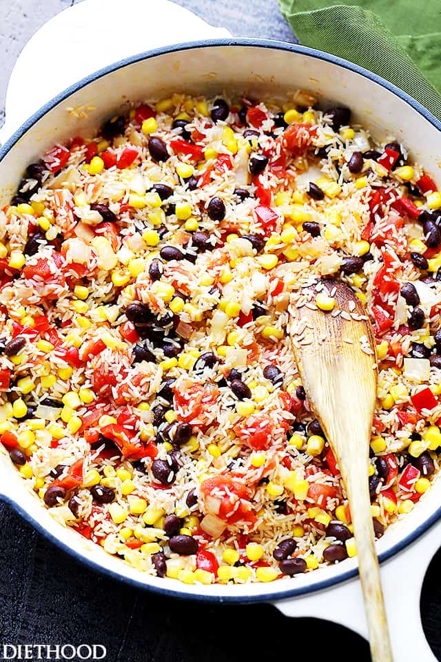 Cooking rice, black beans, tomatoes, corn, and peppers in a white pan and stirring through with a wooden spoon.