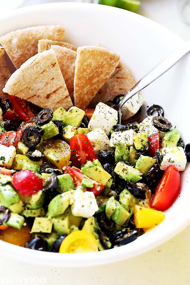 Salad with avocados, olives, and tomatoes layered in a white salad bowl and pita bread wedges arranged around it.