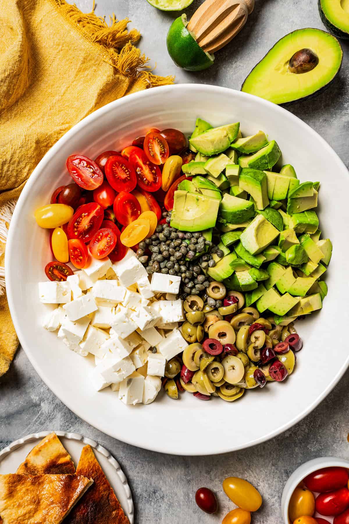 Halved grape tomatoes, cubes of feta cheese, diced avocado, and sliced olives, all arranged in a large salad bowl.