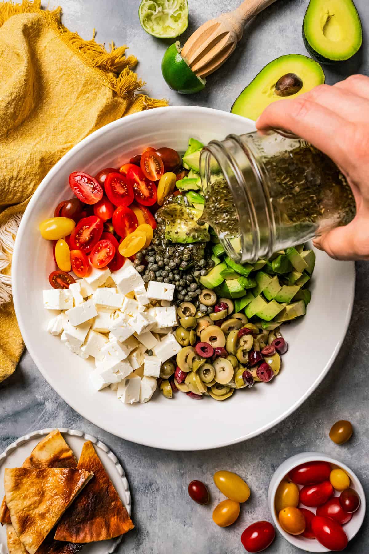 A hand pouring dressing from a jar over avocado salad in a large bowl.