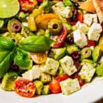 Avocado salad with olives, tomatoes, and feta in a large salad bowl garnished with fresh basil and lime slices.