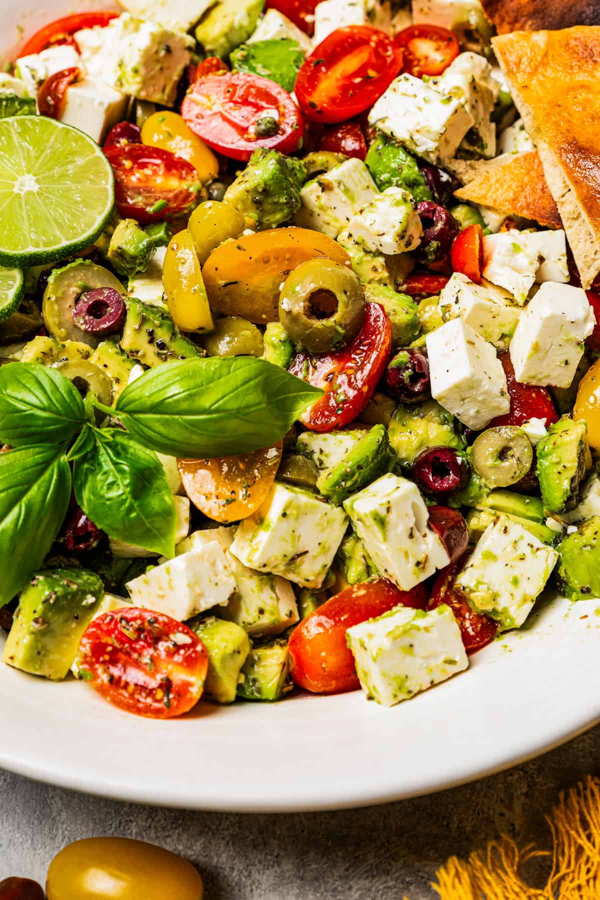 Avocado salad with olives, tomatoes, and feta in a large salad bowl garnished with fresh basil and lime slices.