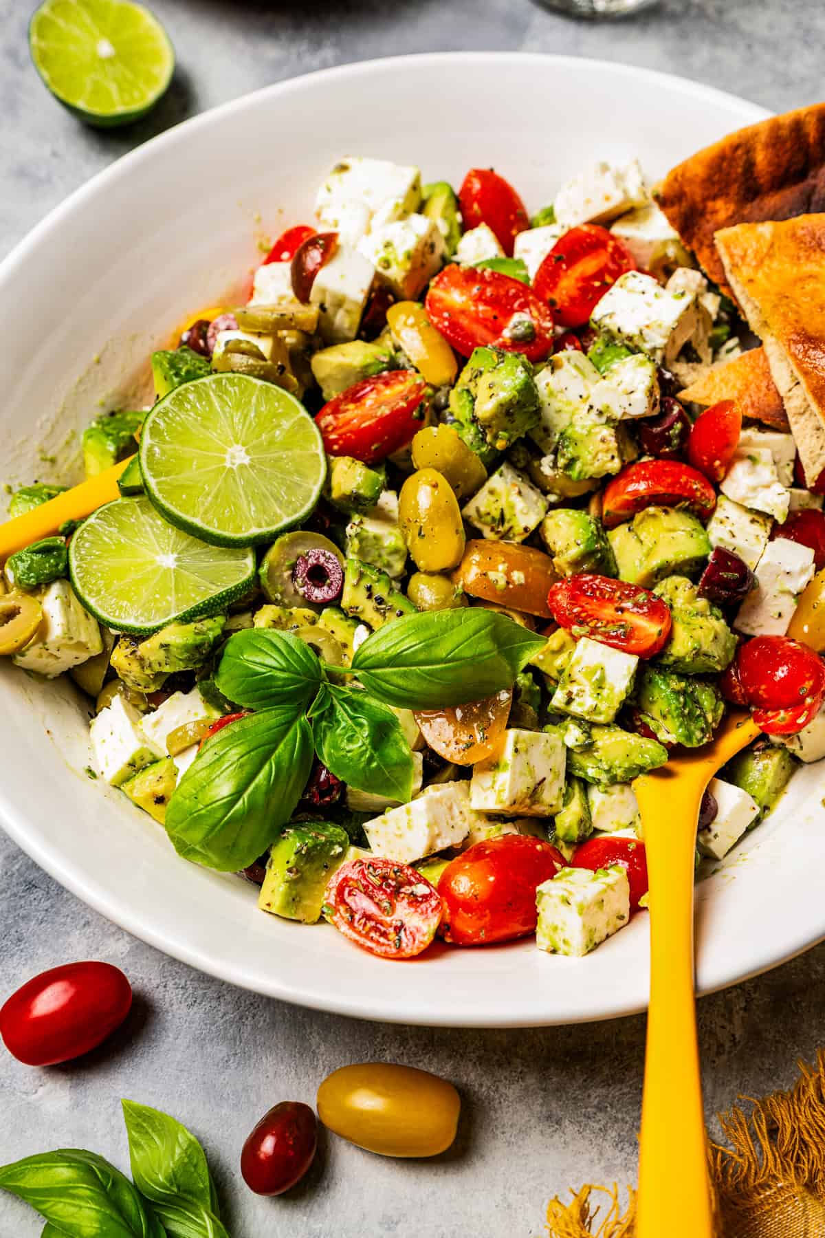 Diced avocados, olives, tomatoes, and feta in a large salad bowl garnished with fresh basil and lime slices.