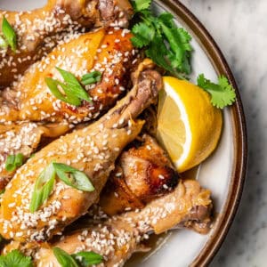 Close up overhead view of crock pot chicken drumsticks garnished with sesame seeds, orange wedges, and green onions on a stoneware plate.