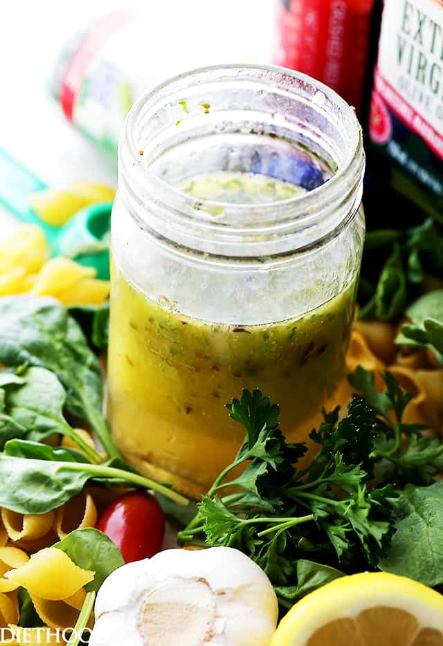 Homemade Italian Salad Dressing in a jar, surrounded by lemons, garlic, fresh greens, and dry pasta.