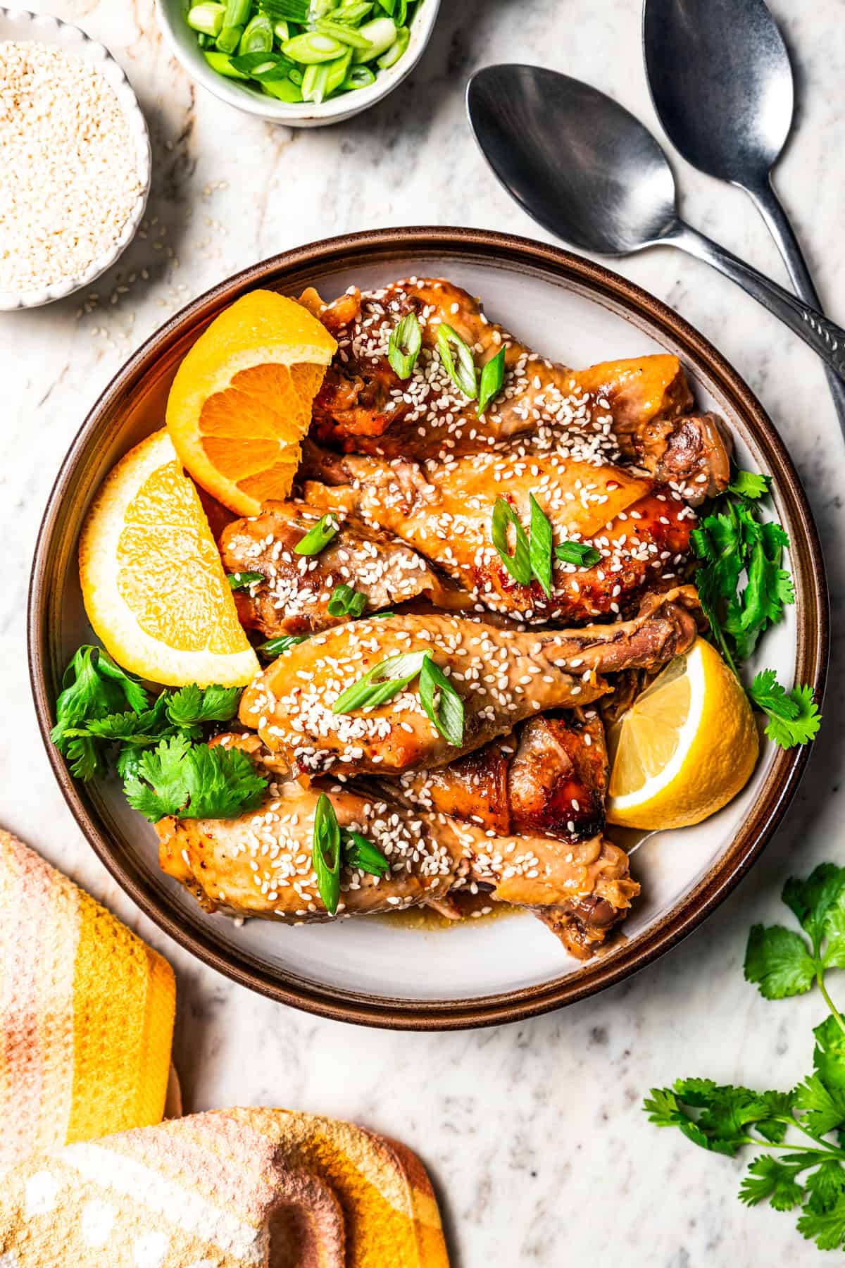 Overhead view of crock pot chicken drumsticks garnished with sesame seeds, orange wedges, and green onions on a stoneware plate.