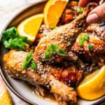 A hand lifting a chicken drumstick from a plate of chicken garnished with orange slices, sesame seeds, and green onions.