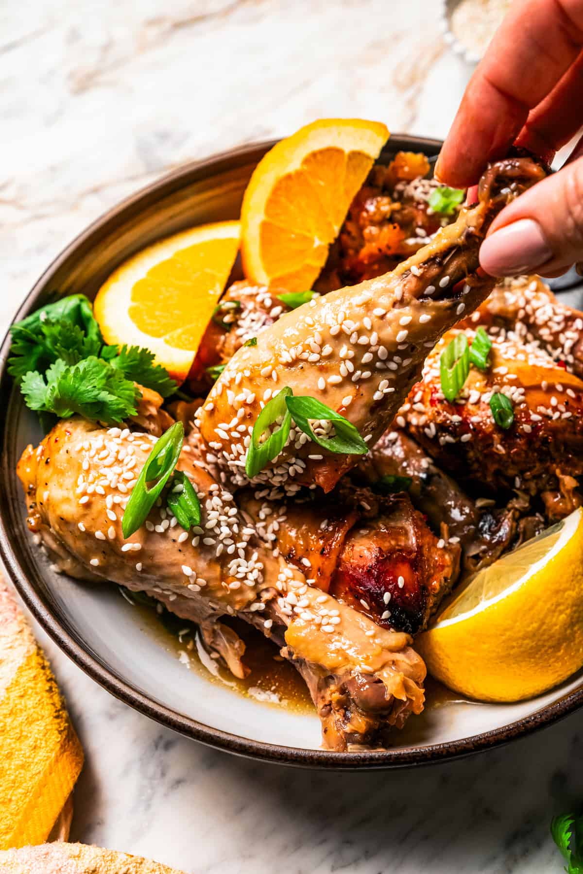 A hand lifting a chicken drumstick from a plate of chicken garnished with orange slices, sesame seeds, and green onions.