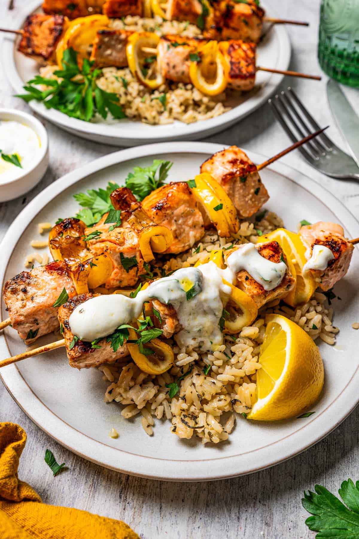 These juicy salmon kabobs with lemon are perfect for grilling! Serve with garlicky yogurt dipping sauce for a delicious and easy summer meal. Two salmon kabobs served over a bed of rice on a plate, topped with garlic sauce and garnished with lemon wedges.