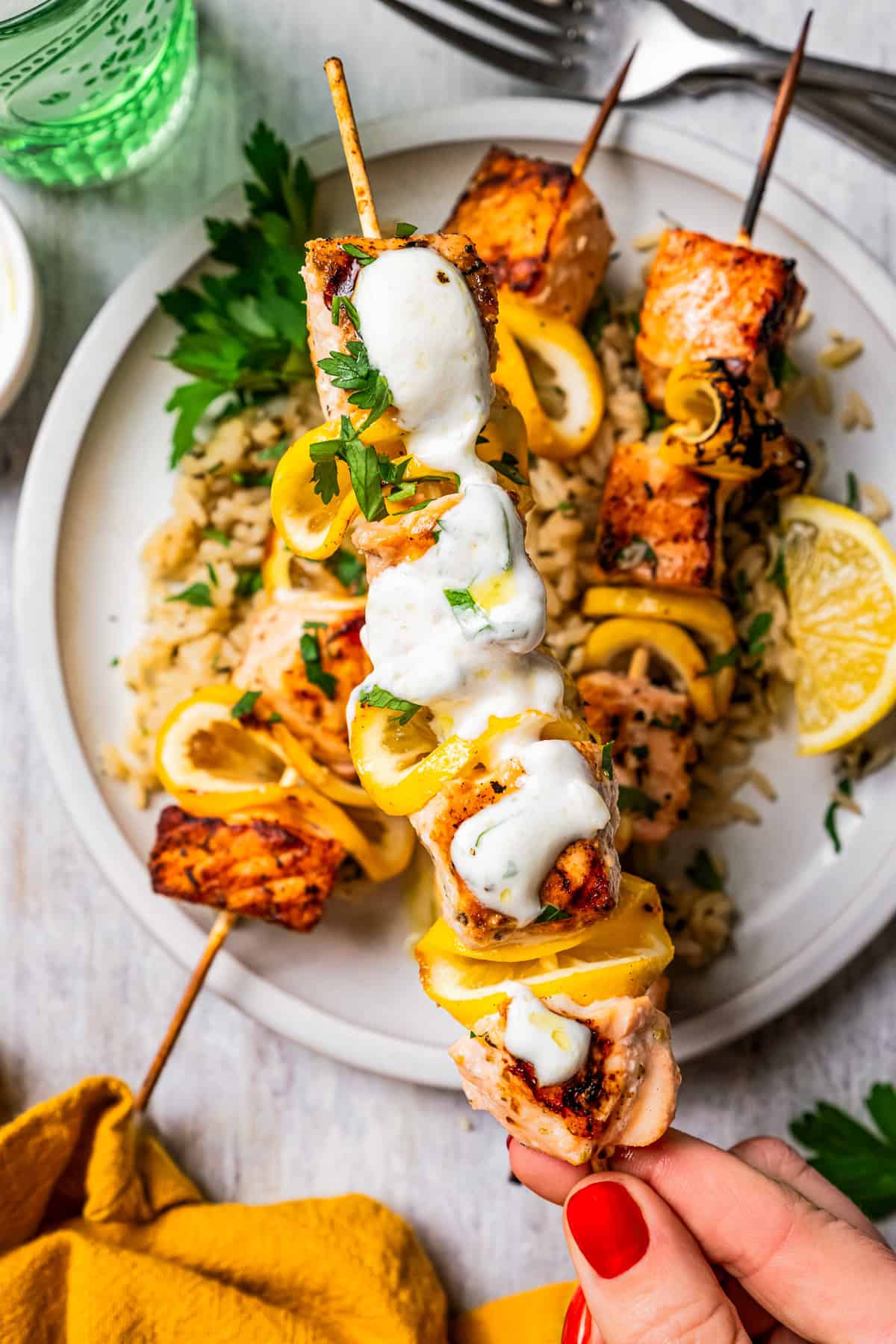 These juicy salmon kabobs with lemon are perfect for grilling! Serve with garlicky yogurt dipping sauce for a delicious and easy summer meal. A hand holding a salmon kabob topped with garlic sauce above a plate.