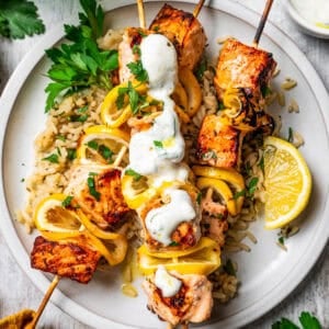 Salmon skewers served over a bed of rice on a plate, topped with garlic sauce and garnished with lemon wedges.