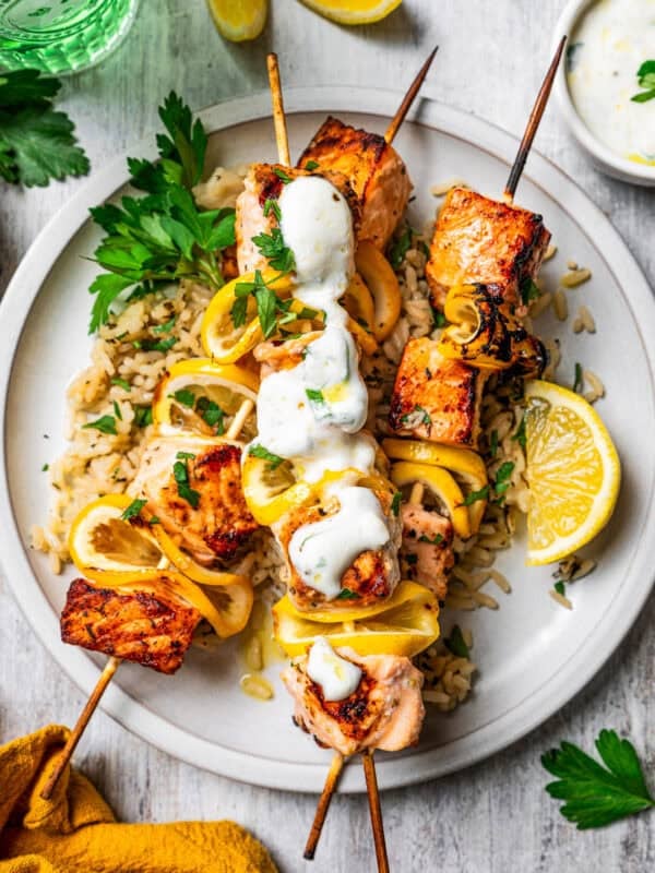 Salmon skewers served over a bed of rice on a plate, topped with garlic sauce and garnished with lemon wedges.