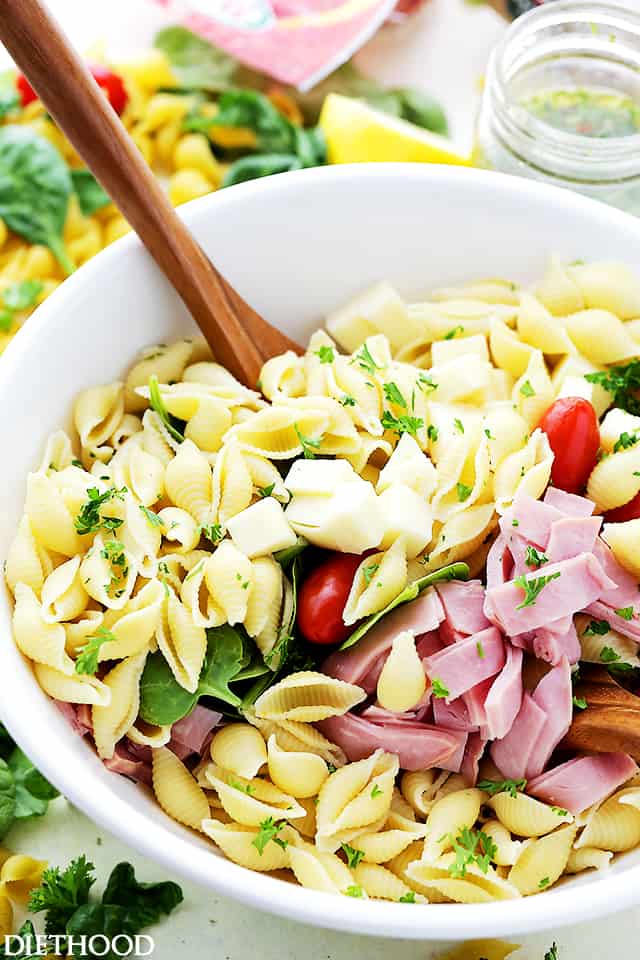 Italian pasta salad ingredients combined in a white bowl with a wooden spoon.