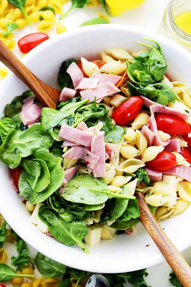 Two wooden spoons resting in a bowl of combined Italian pasta salad ingredients.