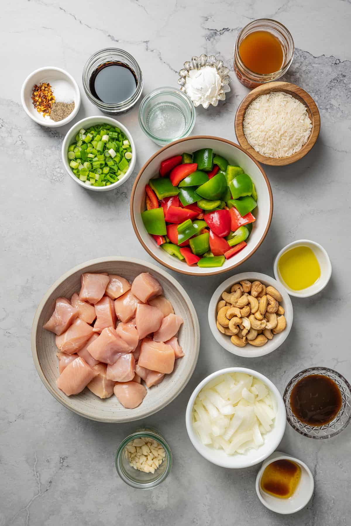 Overhead view of the ingredients needed for cashew chicken: A bowl of raw chicken cubes, a bowl of red and green bell peppers, a bowl of scallions, a bowl of cashews, a bowl of oyster sauce, a bowl of uncooked rice, a bowl of chicken broth, a bowl of cornstarch, a bowl of soy sauce, a bowl of olive oil, a bowl of sesame oil, a bowl of garlic, a bowl of rice vinegar, a bowl of onions, and a bowl of salt, pepper, and chili flakes