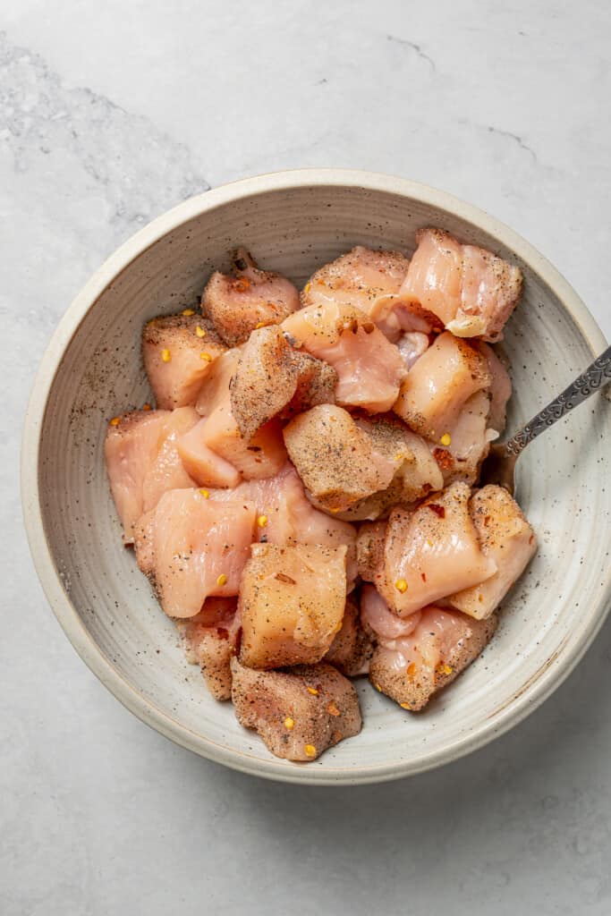 Raw chicken cubes in a mixing bowl with salt, pepper, chili flakes, and a spoon