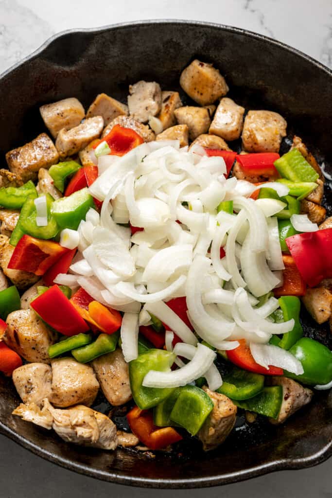 Raw bell peppers and onions on top of cooked chicken chunks in a skillet