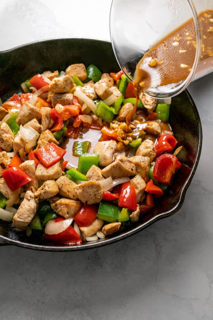A pyrex of chicken broth being poured into a skillet of bell peppers and chicken