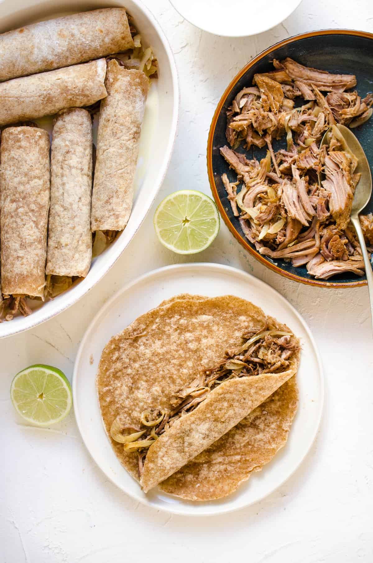 A flour tortilla, partially rolled up with pulled pork filling inside. More rolled tortillas are in a baking dish nearby.
