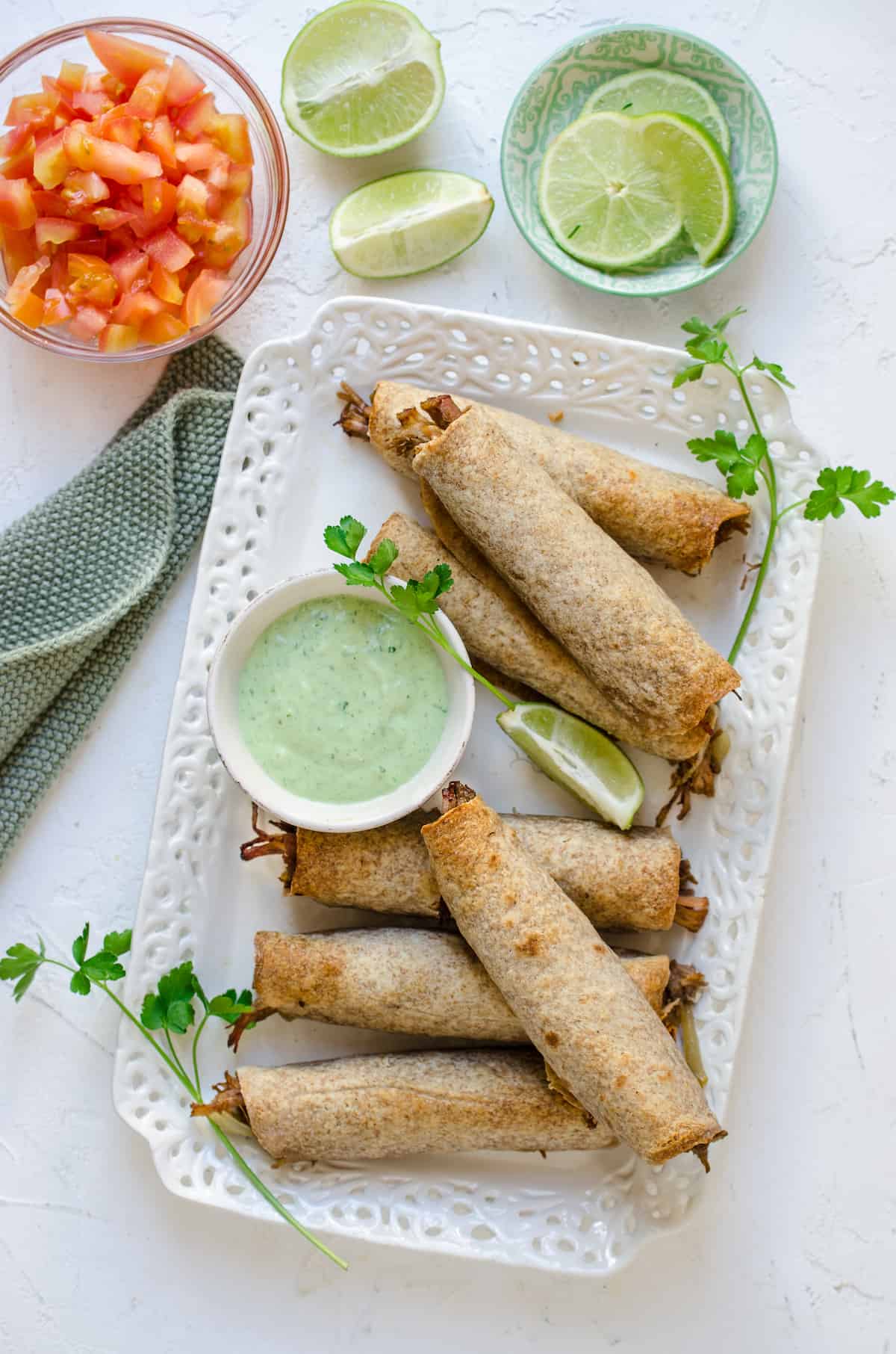 Flautas on a rectangular serving dish, with a white cup of green avocado sauce.