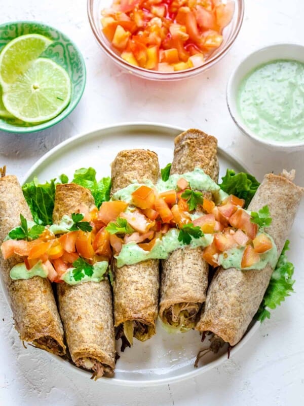 A round white plate with rolled, stuffed flour tortillas, topped with chopped veggies and creamy sauce. Sliced limes, chopped tomatoes, and more sauce are arranged around the plate in little cups.