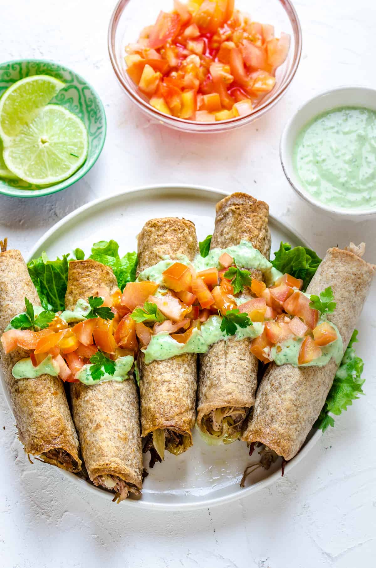 A round white plate with rolled, stuffed flour tortillas, topped with chopped veggies and creamy sauce. Sliced limes, chopped tomatoes, and more sauce are arranged around the plate in little cups.