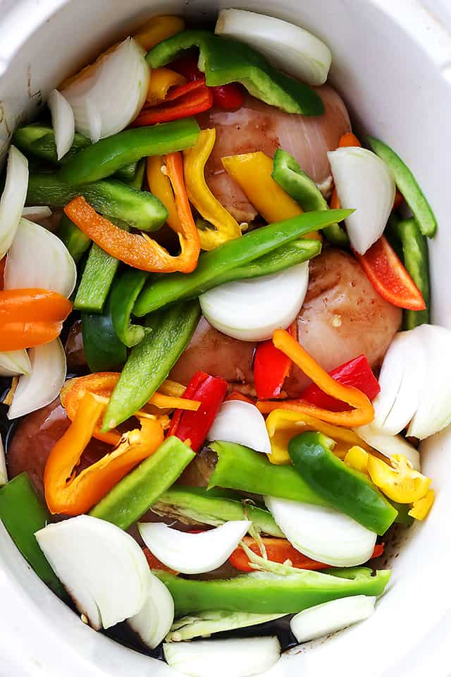 Chopped peppers and onions layered over raw chicken breasts in a crock pot insert.