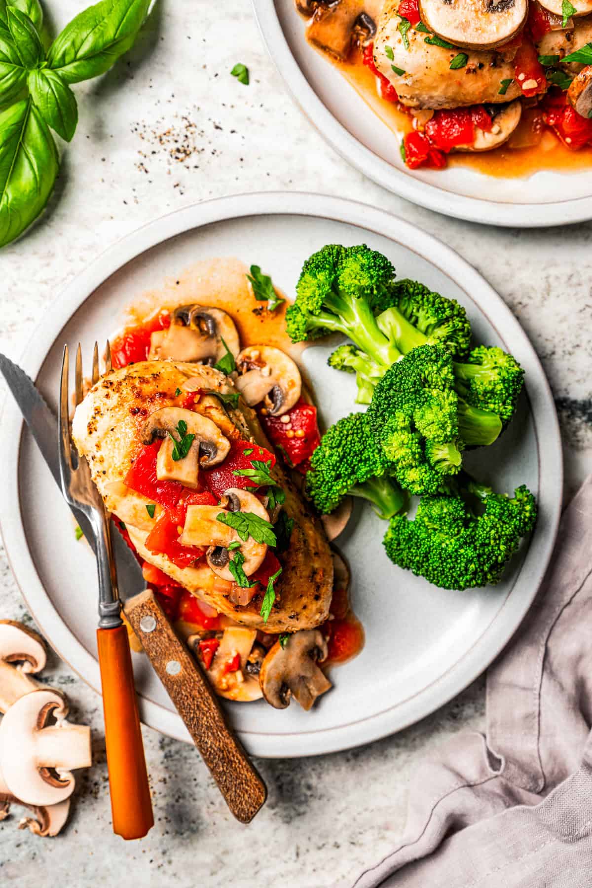 Overhead view of a serving of Chicken Marengo next to a side of steamed broccoli on a plate, next to a fork and knife, with a second plate nearby.