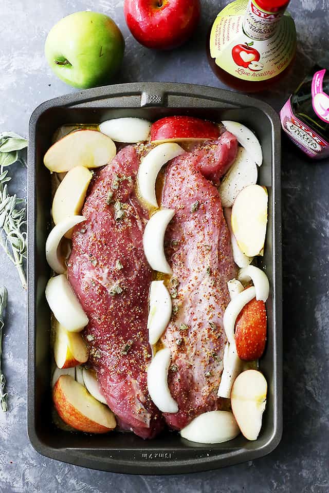 Two cuts of pork tenderloin surrounded by apple and onion wedges in a pan before being cooked