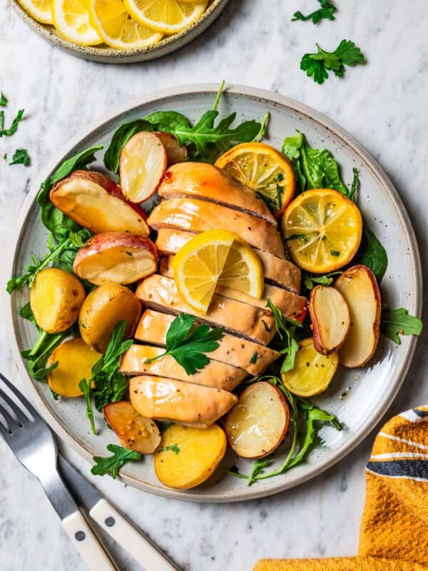 Honey garlic lemon chicken breast cut into slices, surrounded by roasted potatoes and lemon slices on a plate.