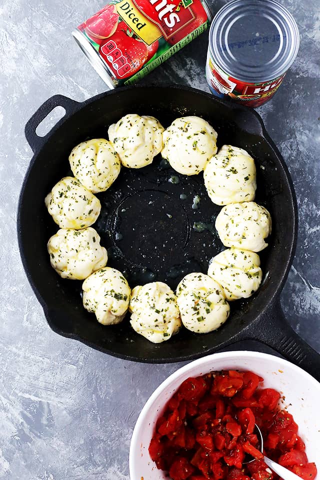 Overhead view of dough balls arranged in a ring in a cast iron skillet next to a bowl of diced tomatoes