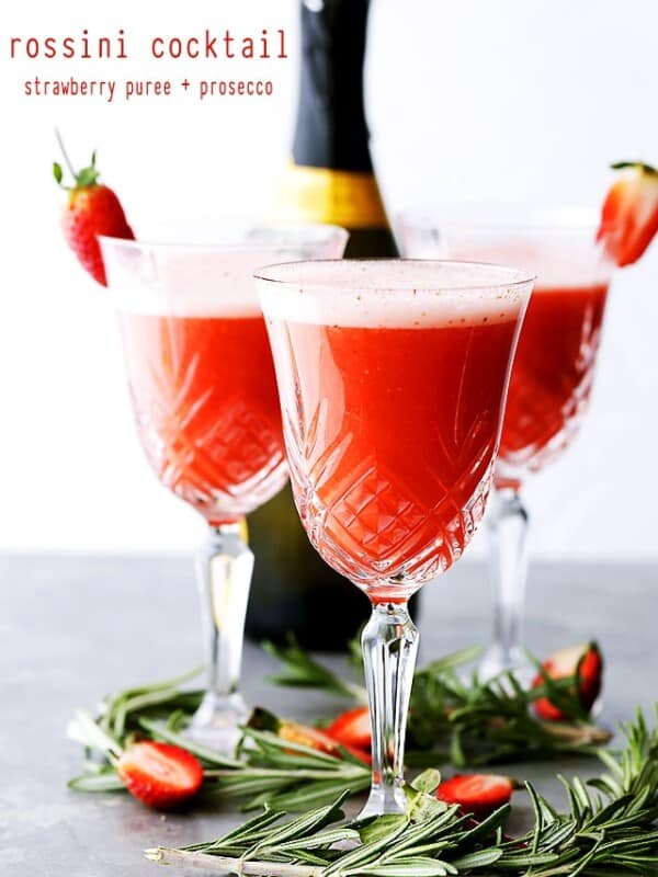 Three Rossini cocktails in crystal flute glasses garnished with strawberries, with a bottle of Prosecco in the background.