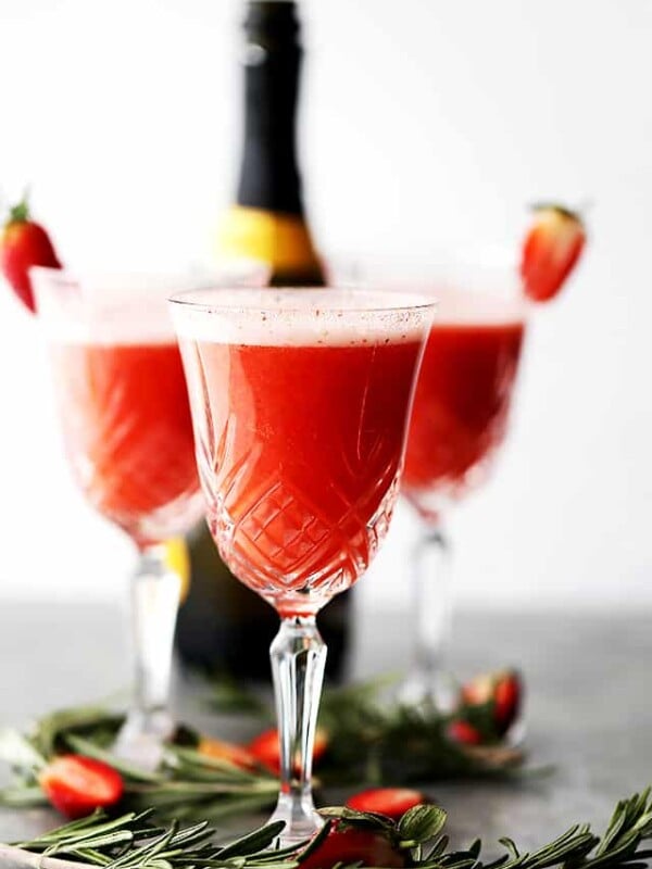 Three Rossini cocktails in crystal flute glasses garnished with strawberries, with a bottle of Prosecco in the background.