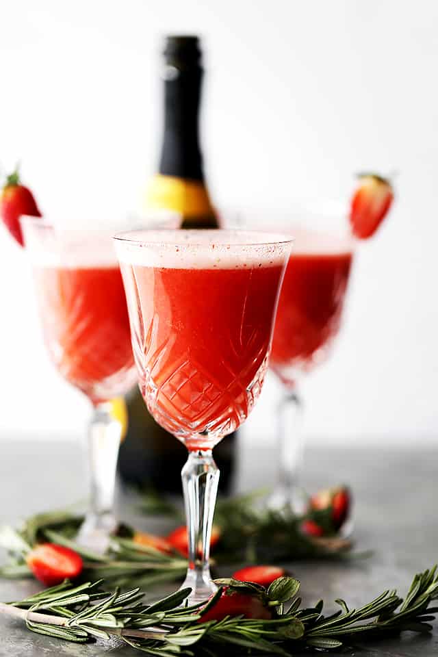 Three Rossini cocktails in crystal flute glasses garnished with strawberries, with a bottle of Prosecco in the background.