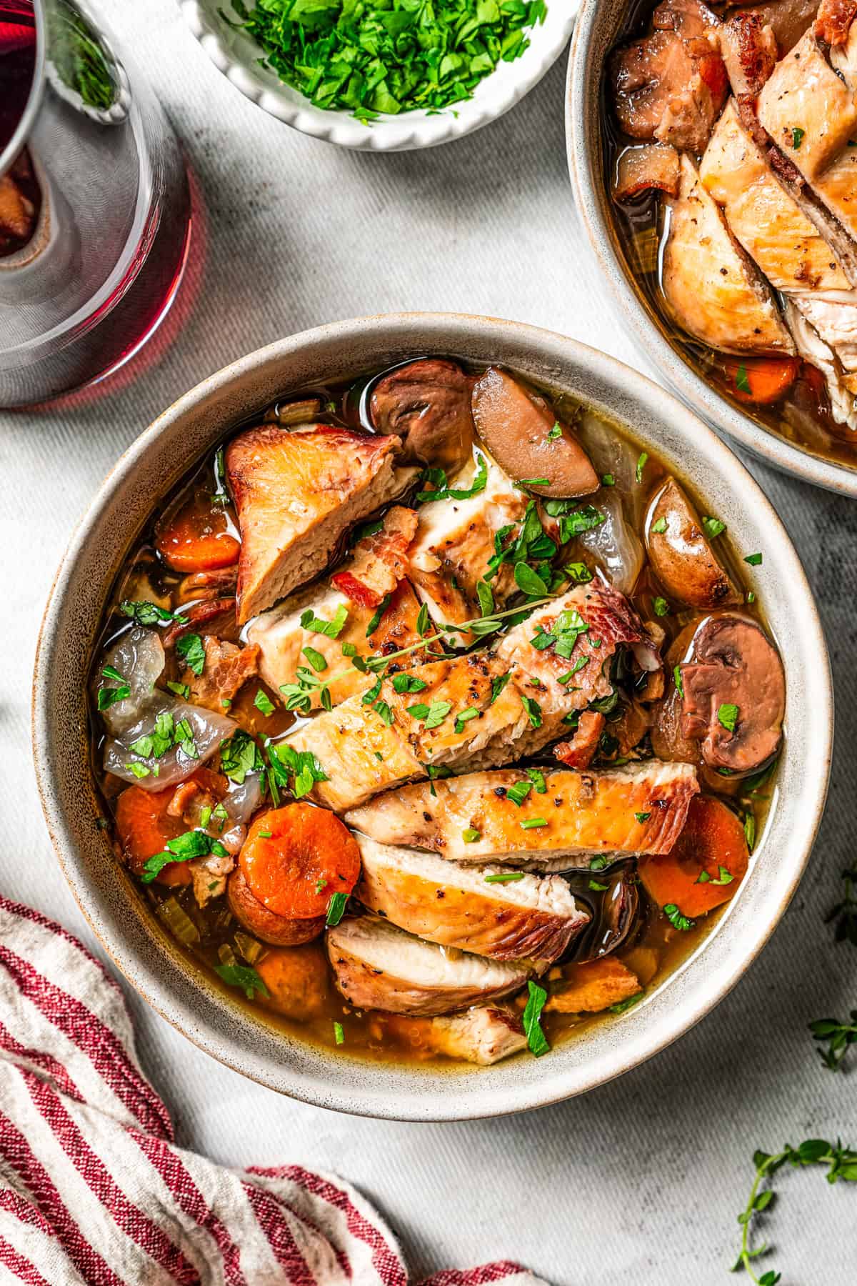 Slow cooker coq au vin served in a white bowl, with a second bowl and a glass of red wine in the background.