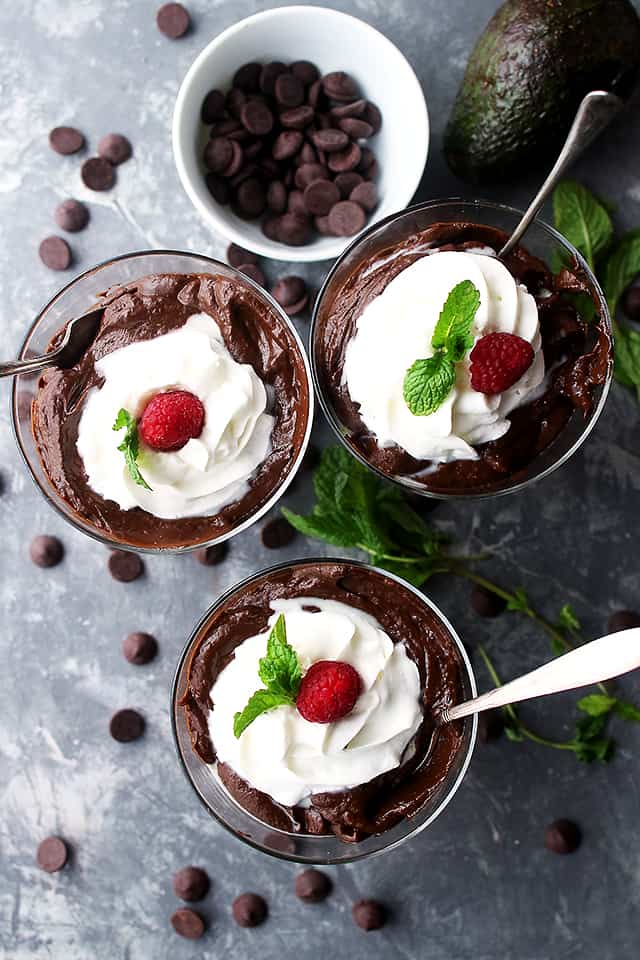 Overhead view of three glasses of avocado chocolate mousse topped with whipped cream, raspberries, and mint leaves, next to a bowl of chocolate chips.