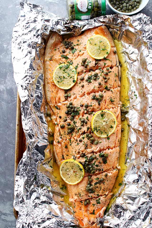 A whole salmon fillet is placed on aluminum foil and topped with lemon slices and capers.