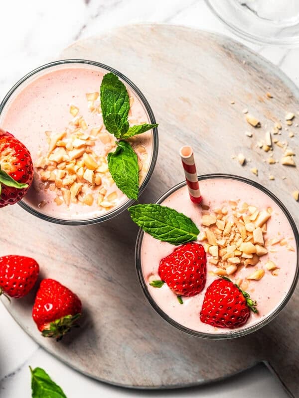 Overhead view of two strawberry protein shakes in glasses garnished with fresh berries, mint leaves, and chopped nuts.