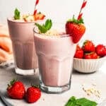 A strawberry protein shake in a glass garnished with fresh berries, mint leaves, and chopped nuts, with a second shake in the background.