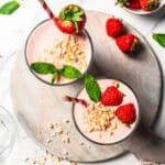 Overhead view of two strawberry protein shakes in glasses garnished with fresh berries, mint leaves, and chopped nuts.