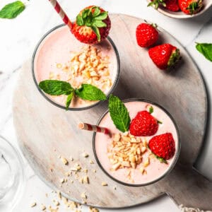 Overhead view of two strawberry protein shakes in glasses garnished with fresh berries, mint leaves, and chopped nuts.