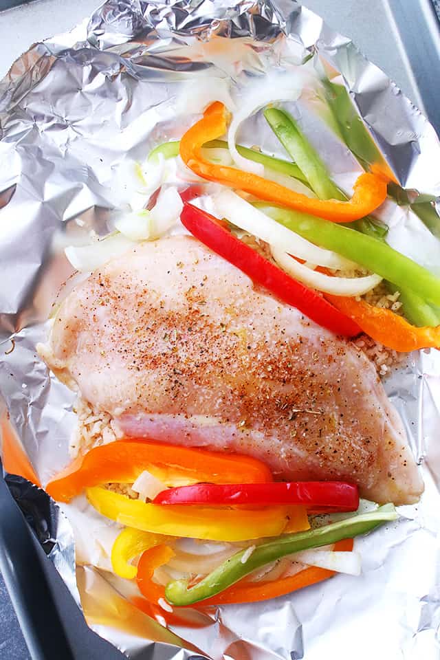Raw chicken breast arranged on a piece of foil with sliced bell peppers and onions placed on either side of the chicken.
