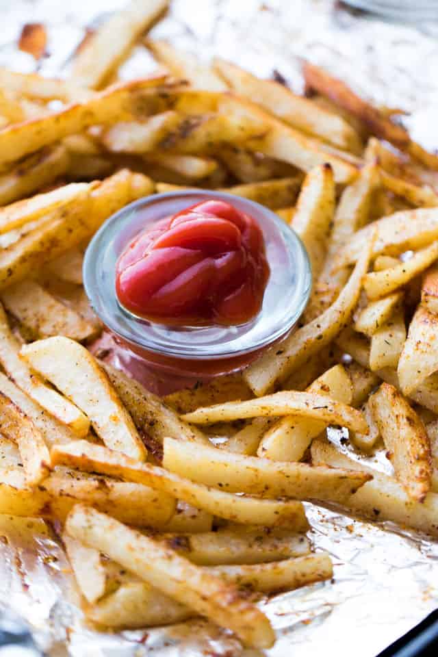 Baked seasoned french fries are arranged on aluminum foil, and a small bowl of ketchup is served with them.