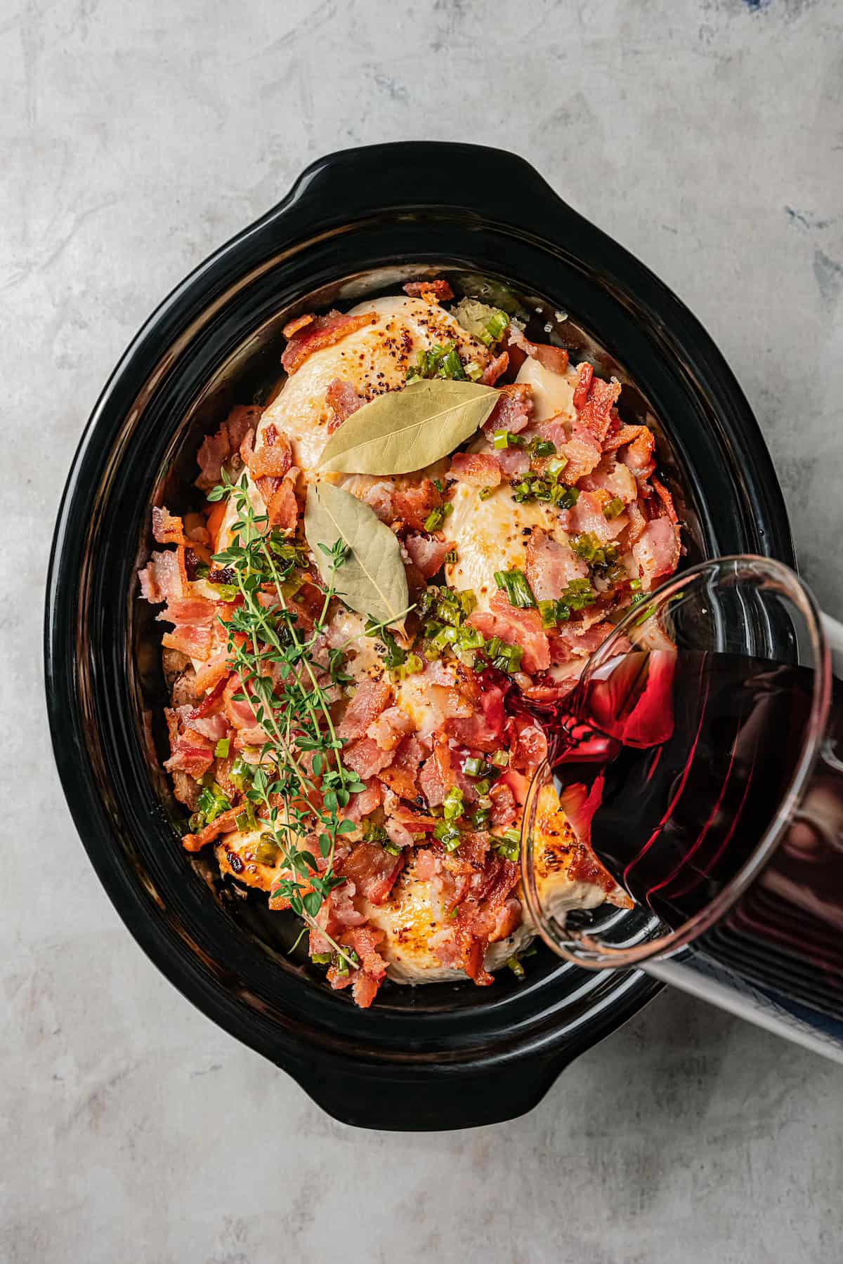 Red wine being poured over coq au vin ingredients inside the slow cooker.