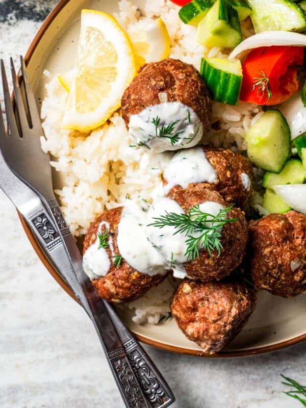 Overhead view of baked chicken meatballs topped with garlic dill yogurt sauce served over rice, next to a side of salad on a plate.