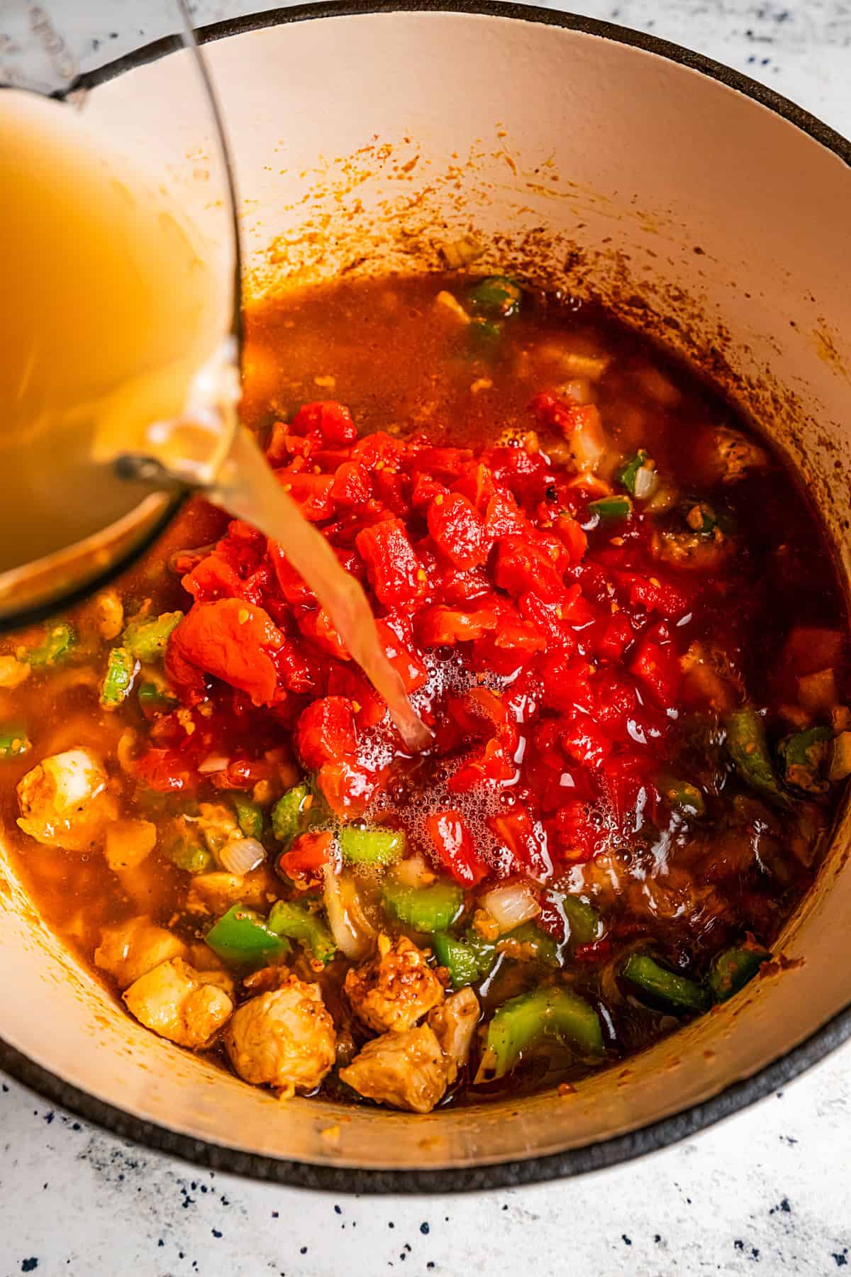 Pouring broth over diced tomatoes, celery, and chicken pieces in a Dutch oven to make Gumbo.